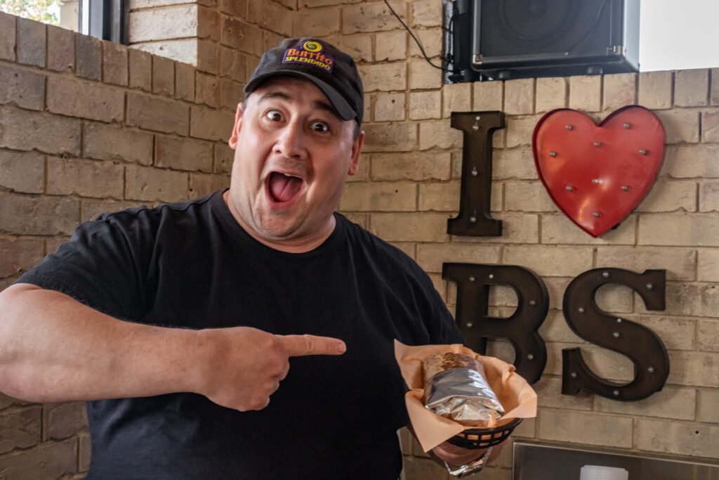 Excited man in a black t-shirt and cap pointing at a wrapped burrito in a basket against a brick wall background.